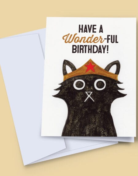 A 7x5 inch birthday card featuring an ink and salt black cat illustration wearing Wonder Woman's head dress