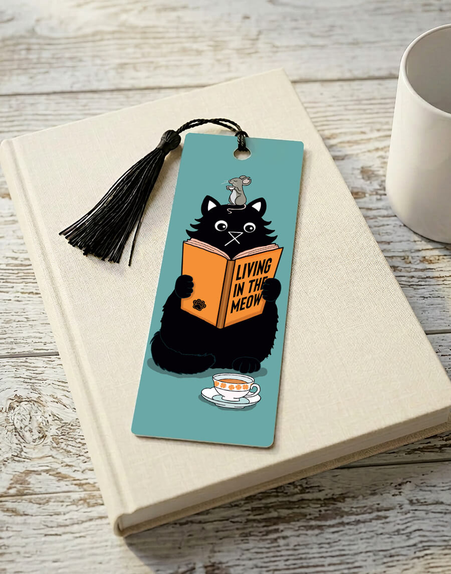 A bookmark featuring a black cartoon cat reading a book titled "Living in the Meow" with a mouse sitting on its head enjoying a cup of tea